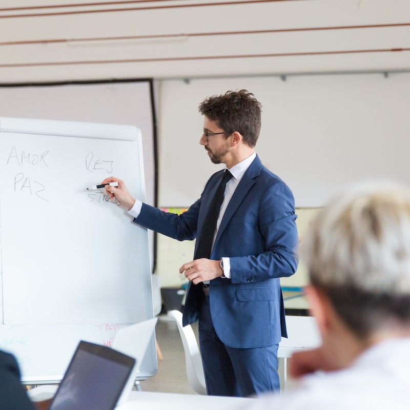 Confident speaker in eyeglasses talking near whiteboard. Group of workers discussing presentation of new project at briefing. Business meeting concept